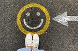 A smiley face in chalk on the ground beneath a woman’s feet, with an arrow pointing at it.