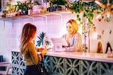 A woman at a shoppe and a Receptionist at a computer at the counter. Many plants on a high shelf