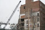 A wrecking ball on a crane swinging towards an old partially-wrecked brick building.