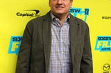Ted Sarandos standing in front of a primarily yellow step and repeat at SXSW in 2016, he is wearing a brown/greenish jacket, a plaid shirt and blue jeans.