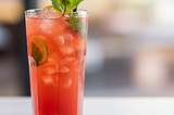 A mocktail in a tall glass. The drink is reddish and has a couple of leaves at the top, inaddition to a straw.