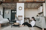 Woman working home home in living room, laptop on her lap, leaning against a grey couch. The apartment is decorated with boho furniture and fairy lights.