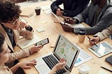 Grupo de pessoas em uma reunião ao redor de uma mesa de madeira clara. Na mesa, há diversos dispositivos tecnológicos, como laptops, tablets e smartphones, exibindo gráficos e dados. Há também papéis, canecas de café, e canetas espalhadas pela mesa.