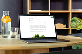 A imagem mostra um laptop aberto sobre uma mesa redonda de madeira clara. Na tela do laptop, há um texto em português com o título “Se a vida lhe der limões…”. Ao lado do laptop, encontra-se uma jarra de vidro contendo água e fatias de laranja, além de um copo vazio. Ao fundo, há uma estante de madeira com prateleiras, decorada com algumas plantas verdes, conferindo um ar acolhedor e organizado ao ambiente.