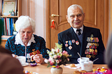 Photo of an eldery lady and eldery man sitting at a table with coffee. The old man is a war veteran wearing a suit ocvered in medals