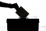 A black on white image of a hand placing a vote into a ballot box.