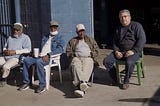 Four men sit in mismatched plastic chairs outside a blue brick auto shop.