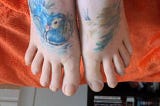 Photo of a pair of white feet on an orange blanket. The left foot has a tattoo of a Mandarin duck among some reeds, mostly done in blue. The right foot has a tattoo of a lake and more reeds. There is a bookcase full of books in the background.