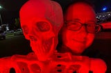 The author is standing behind a skeleton under a red light. He has very short hair and glasses. He’s smiling. The skeleton’s eyes are decorated with glitter.
