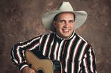 Headshot of Garth Brooks in his early 30s. He is holding a guitar and smiling at the camera, wearing a striped Western shirt and white cowboy hat.