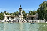 A semicircular set of columns surround a main statue on a high pedestal. The statue is a horse and rider, and the pedestal has steps at the bottom, leading down to a large man-made pond. There are small rowboats in the pond with people rowing. The sky is bright and blue, and there are trees surrounding the scene.