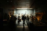 A softly lit museum gallery with glass display cases holding artifacts. Several visitors, shown in silhouette, examine objects up close or chat nearby. Large statues and pottery line the walls, illuminated by spotlights. The modern space features wood-paneled ceilings and a polished floor that reflects the exhibits, creating a sense of depth and quiet curiosity.