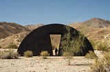A giant, half-moon, black sculpture sits in the desert landscape. Behind it are sandy hillsides. At its center is an angular cutout, a doorway with the silhouette of a person standing inside.