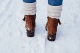 Close-up of a person’s legs wearing brown suede boots, cream knit socks, and blue jeans, walking through fresh snow.