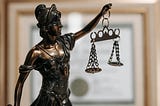 a close-up photo of a bronze figurine of Lady Justice from the waist up, with a blurred-out academic degree in a picture frame in the background