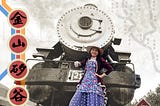 Photo of Alfred in a prairie dress and apron, standing in front of an old steam locomotive. In the background are a street map made up of streets from various Chinatowns and Asian American neighborhoods and a railway map of California cities with Chinese names.