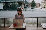 A women on her mac laptop facing the camera, sitting in a open patio cafe looking on to the intersection