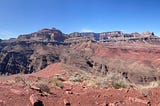 Panorama photo of the Grand Canyon. Blue sky above red rocky soil with a river at the bottom of a deep canyon. There is a shadow of a hiker on the right side.