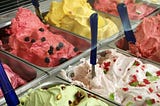 A traditional gelato display with several different flavours in metal trays with spatulas stuck into the,