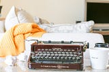 Corona typewriter on table with crumpled pieces of paper beside it, a white sofa and yellow blanket behind, and a coffee mug.