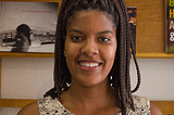 Juliana é uma mulher negra, e está com os cabelos castanhos trançados e presos. Ela está sorrindo e vestindo uma roupa regata de renda branca com detalhes pretos. Ao fundo, há uma prateleira de madeira com quadros