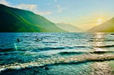 A shoreline with waves rolling in. Children play in the water further out. The sun sets behind hills in the distance