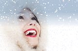 A young woman wearing a fur-trimmed hood looks up into falling snow, laughing.