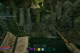 A first-person perspective camera view of the game Avowed, showing a lush interior cave, with a small pond just ahead and some mushrooms. The player character holds up a wand a magical book in their hands.