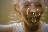 A photograph of a small child under the water with her eyes squeezed shut and surrounded by the air bubbles she is blowing out.