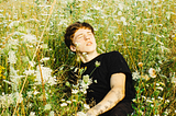 A young white man with short, slightly-ginger hair, wearing a black T-shirt and sitting and leaning back on his right arm in a field of tall green and amber grass and while flowers. It’s a sunny day, his eyes and closed and his face is turned towards the sun.