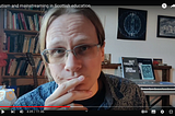 A video still of the author, a white person with light hair and glasses, pausing in mid-flow with a sceptical expression. Their room is full of books, art and musical instruments.