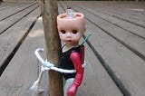 Photo of an eerie-looking little doll wearing a onesie and missing the top half of her skull, tied to a stake with a tiny firecracker strapped to her back.