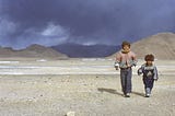 Two children dressed in warm clothes walking across a bleak landscape.
