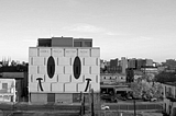 A black and white image of a building with a smiley face mural. Buildings and a street intersection in the background.