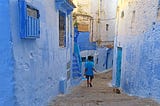 Man in a blue tshirt walks down a narrow street with blue, plastered buildings on either side. Blue doors, blue windows, blue stairs.