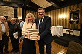 A smartly dressed middle-aged man and woman stand holding a pile of books at a book-signing event in a traditional hotel function room