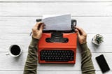 person’s hands removing paper from an orange typewriter on a white surface with a cup on left, phone and plant on right