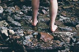 Woman standing barefoot on rocks with water flowing past.