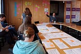 The team sit around a table with papers on as Tom Steinberg gestures to a wall of post it notes clustered together.