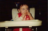 a baby sits in a retro-looking highchair with a party hat on and she’s biting into an orange segment with apparent trepidation