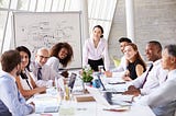 Asian Businesswoman Leading Meeting At Boardroom Table with other leaders, colleagues, and or peers of mixed races white, South-Asian, and Black all of different ages and work experience.