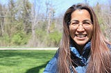 Photo of candidate Maria Peterson, running for IL House of Representatives, District 52. Maria is wearing a blue jacket, standing outside with green grass and trees in the background.