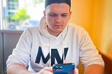 Isaac Mashman wearing a backward hat and white Mashman Ventures hoodie, sitting in front of an orange wall, looking at his phone.