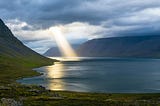 Light streams through clouds onto a large body of water surrounded by hills.