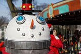 sculpture shows a robot with a silver neck and a white, dome-shaped head. the head is adorned with a carrot nose and a top hat. vaguely electronic features, including silver spheres and blue lights, give the impression of a mouth and eyes. the overall effect is of a robot that resembles a snowman.