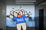 This image shows an illustration of a cartoon koala with an open mouth and closed eyes, wearing a blue shirt with the word “BOSS” on it. The bear appears to be in mid tantrum, with arms stretched wide in an assertive gesture. The cartoon koala is superimposed on a real-life photograph of an elevator lobby.