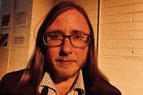 The author is standing in front of a white brick wall wearing a black suit and a pinstriped shirt. He’s wearing glasses and a serious face. He has long brown hair, a beard and a mustache.