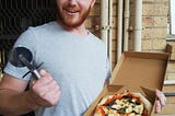 A photo of the author in a grey T-shirt, a small pizza in his left hand and pizza slicer in his right hand. He’s standing next to a small wooden-slatted table, and in front of a mesh-screen door and a tan brick wall with pipes running vertically and horizontally along it. He has an orange beard and short, light orange hair.