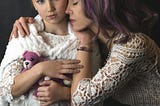 Mother and daughter, both dressed in white lace. The girl has small purple stuffed bear and a purple hair tie, the mother has purple highlights in her hair.