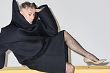 Alan Cumming in a black robe and high heels, posing suggestively for the camera